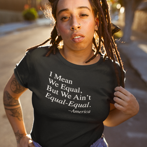 A woman wearing a black ROCKaBLOCK statement t-shirt with the quote "I mean we equa, but we ain't equal-equal' on it, standing outdoors.