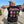Load image into Gallery viewer, Street-style photo of a man wearing a black satin ROCKaBLOCK “Freedom Fighters” varsity jacket with illustrated patches of historic leaders, captured on a sunny city sidewalk.
