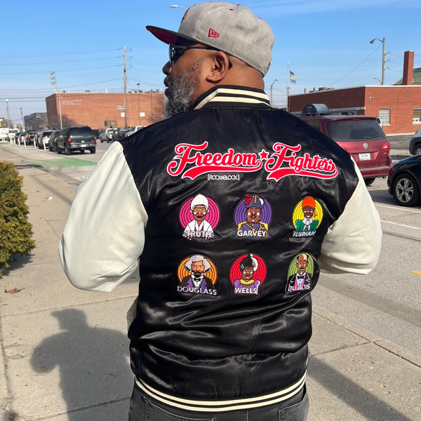Street-style photo of a man wearing a black satin ROCKaBLOCK “Freedom Fighters” varsity jacket with illustrated patches of historic leaders, captured on a sunny city sidewalk.