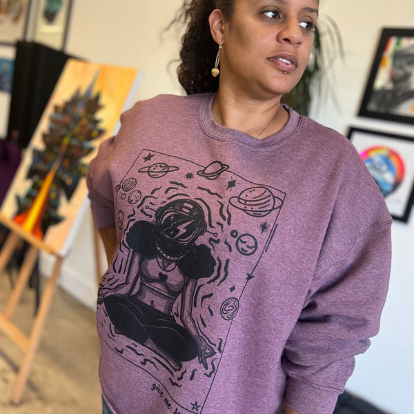 A woman stands in an art-filled studio wearing the ROCKaBLOCK “Give Me Space” raw hem crewneck sweatshirt in mauve, featuring a bold black illustration of a meditating woman in an astronaut helmet surrounded by planets and stars.