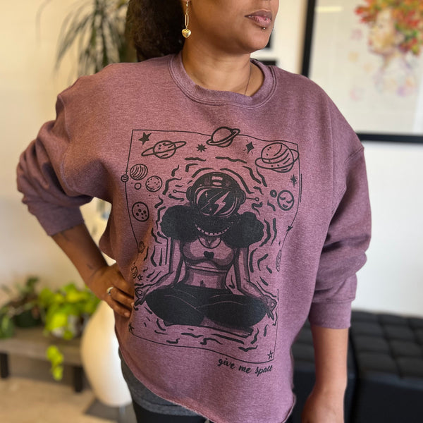 A woman stands in an art-filled studio wearing the ROCKaBLOCK “Give Me Space” raw hem crewneck sweatshirt in mauve, featuring a bold black illustration of a meditating woman in an astronaut helmet surrounded by planets and stars.