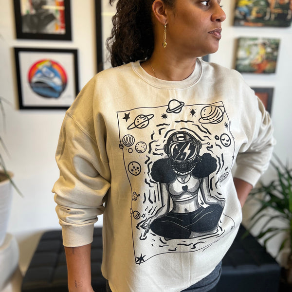 A woman stands in an art-filled studio wearing the ROCKaBLOCK “Give Me Space” raw hem crewneck sweatshirt in tan, featuring a bold black illustration of a meditating woman in an astronaut helmet surrounded by planets and stars.