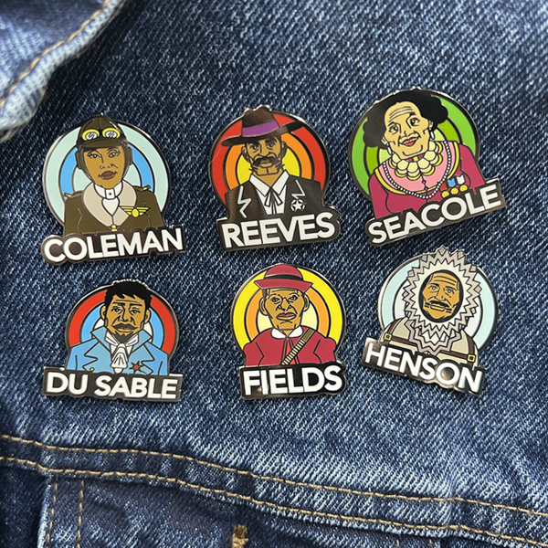 A collection of ROCKaBLOCK hard enamel Pioneers pins with characters including Bessie Coleman, Bass Reeves, 'Stagecoach' Mary Fields, Jean Baptiste Point Du Sable, Mary Seacole, and Matthew Henson, secured on a denim fabric. Highlighting Black history.