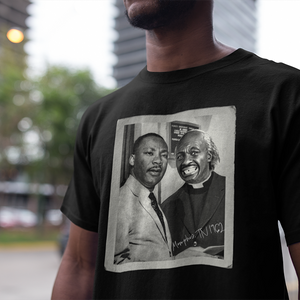 Man wearing a black graphic t-shirt featuring a vintage-style portrait of Martin Luther King Jr. alongside Clarence the Barber, set against a softly blurred urban street background.