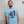 Load image into Gallery viewer, A man with a beard and arm tattoos wearing a light blue t-shirt with a graphic of a boombox and cassette tapes, standing against a textured concrete wall.
