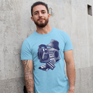 A man with a beard and arm tattoos wearing a light blue t-shirt with a graphic of a boombox and cassette tapes, standing against a textured concrete wall.