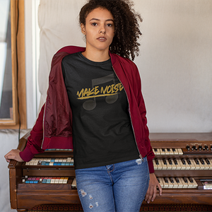 A woman stands in front of a vintage organ wearing a black graphic t-shirt with a yellow and gray 'MAKE NOISE' design, a maroon bomber jacket, and blue jeans.